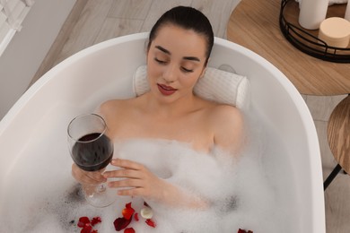 Photo of Woman with glass of wine taking bath in tub with foam and rose petals indoors, above view