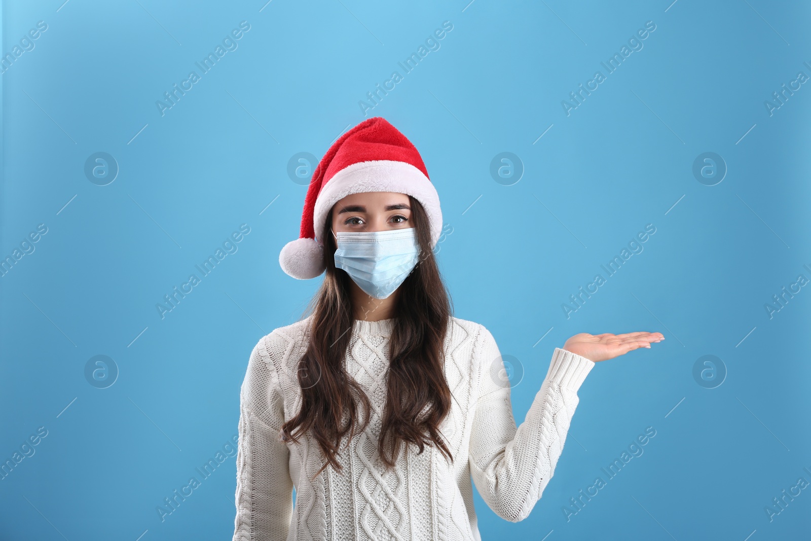 Photo of Beautiful woman wearing Santa Claus hat and medical mask on light blue background