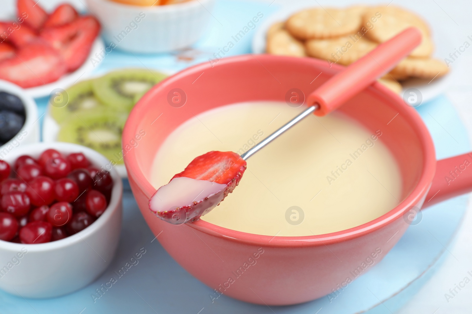 Photo of Fork with strawberry dipped into white chocolate fondue and ceramic pot on table