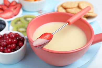 Photo of Fork with strawberry dipped into white chocolate fondue and ceramic pot on table