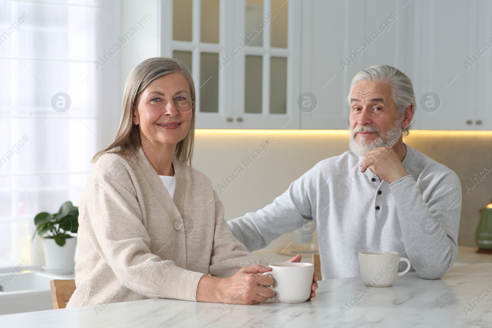 Photo of Affectionate senior couple with cups of drink at white marble table in kitchen