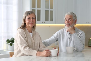 Photo of Affectionate senior couple with cups of drink at white marble table in kitchen