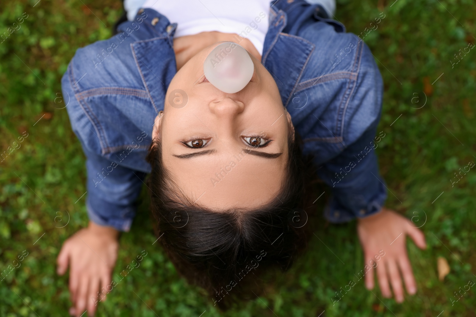 Photo of Beautiful young woman blowing chewing gum on green grass outdoors, top view