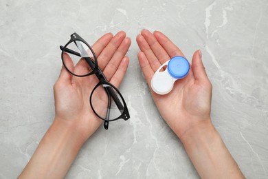 Photo of Woman holding case with contact lenses and glasses at light grey marble table, top view
