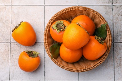 Delicious ripe juicy persimmons in wicker basket on tiled surface, flat lay