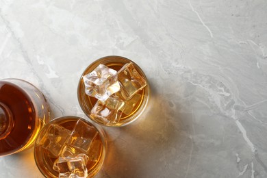 Whiskey with ice cubes in glasses and bottle on light grey textured table, flat lay. Space for text