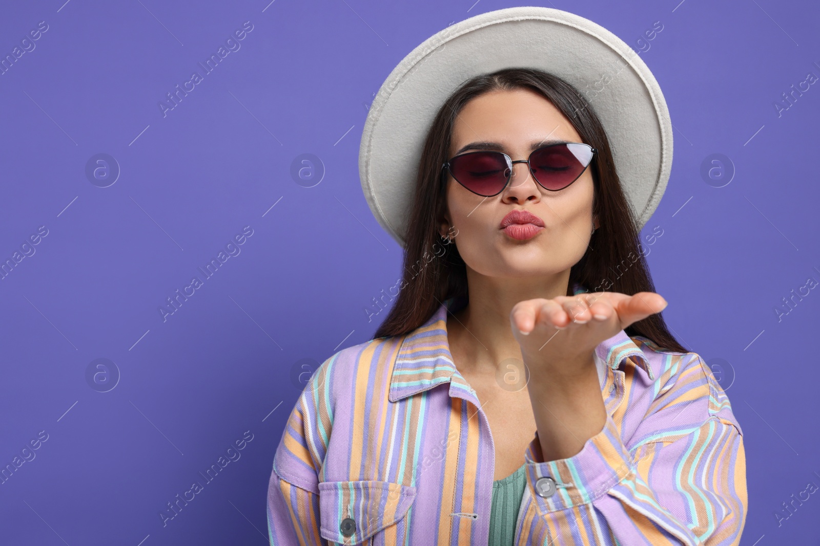 Photo of Beautiful young woman with stylish hat blowing kiss on purple background. Space for text