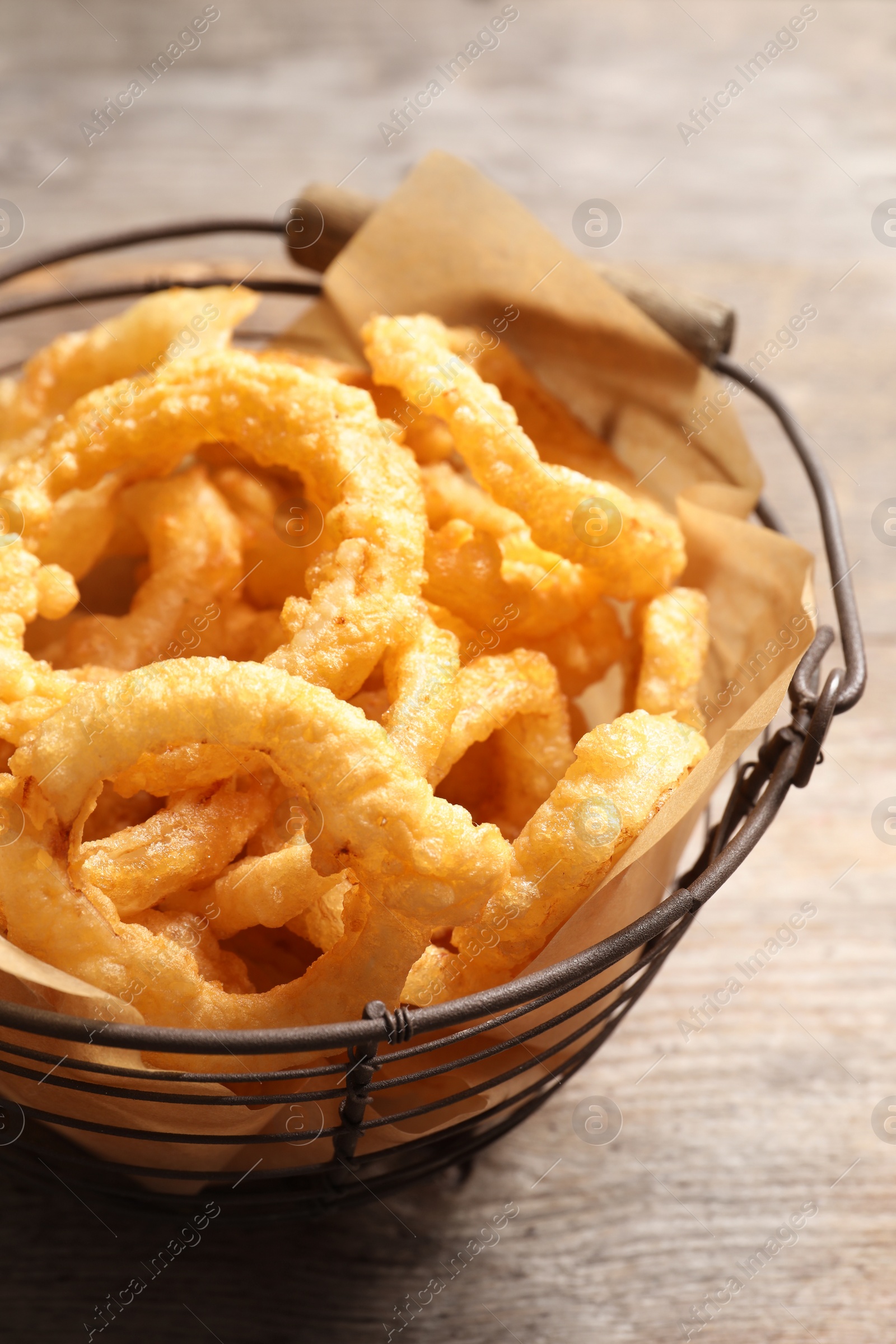 Photo of Homemade crunchy fried onion rings in wire basket on wooden background, closeup