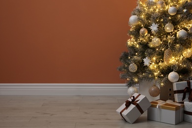 Photo of Beautiful decorated Christmas tree with glowing fairy lights and presents near orange wall indoors. Space for text