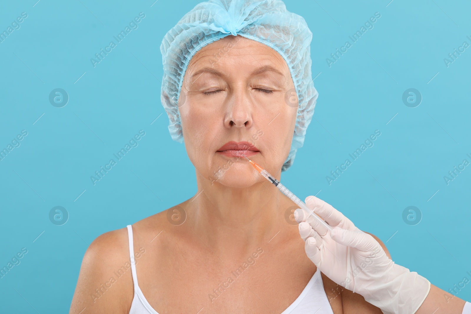Photo of Doctor giving lips injection to senior woman on light blue background. Cosmetic surgery