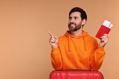 Smiling man with passport, suitcase and tickets pointing at something on beige background. Space for text