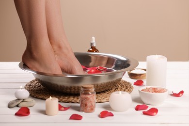 Photo of Woman soaking her feet in bowl with water and rose petals on white wooden surface, closeup. Pedicure procedure