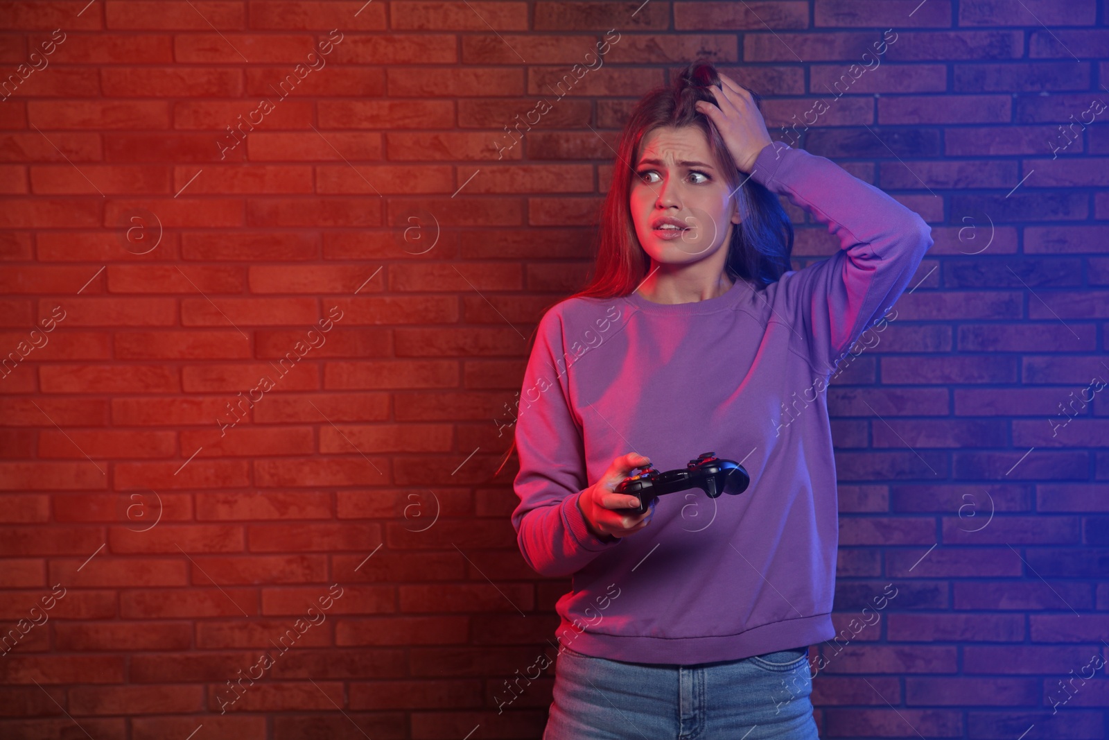Photo of Emotional young woman playing video games with controller near brick wall. Space for text