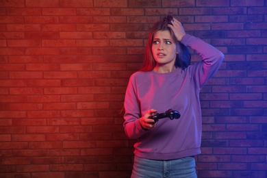 Emotional young woman playing video games with controller near brick wall. Space for text
