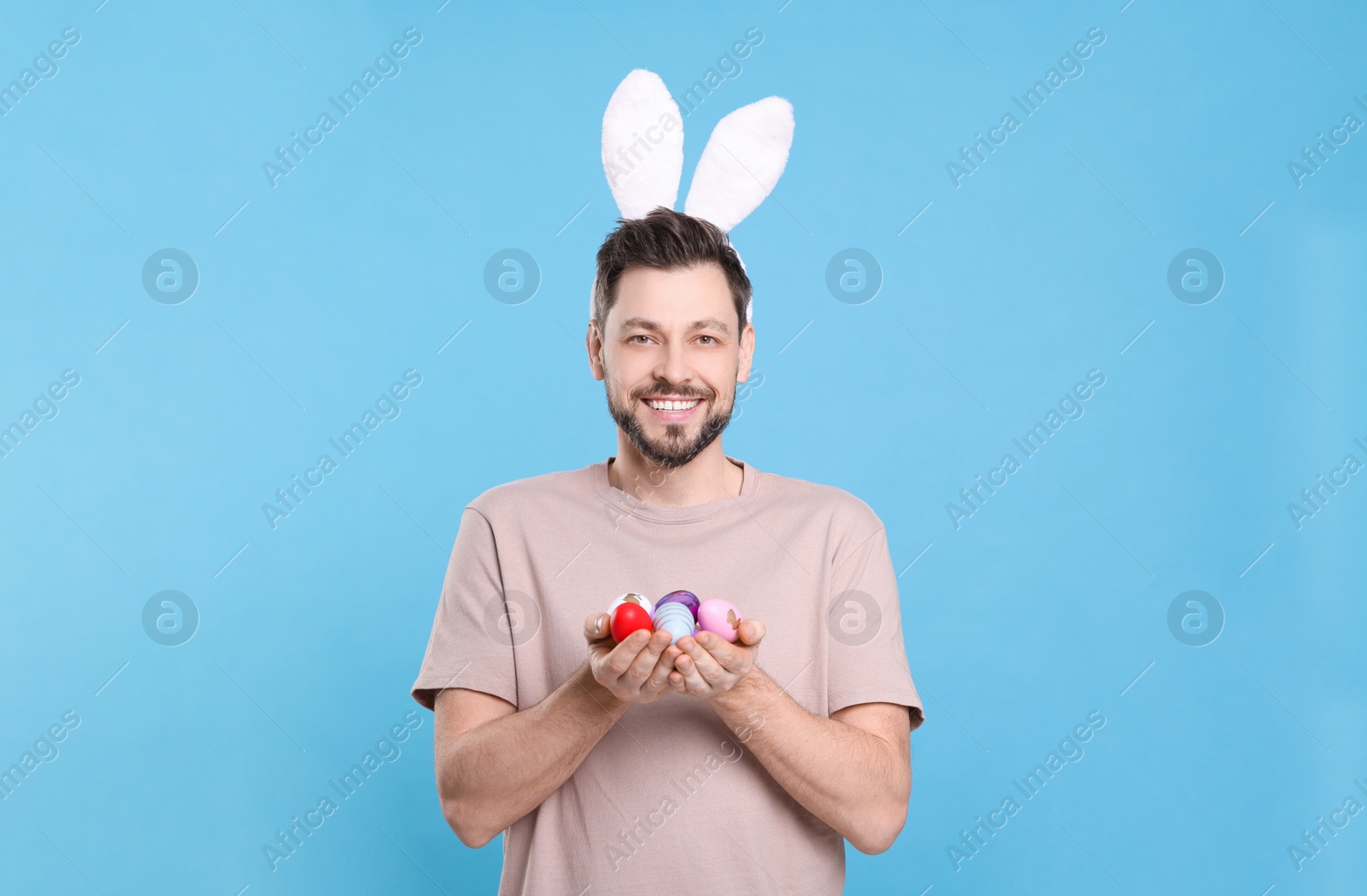 Photo of Happy man in bunny ears headband holding painted Easter eggs on turquoise background