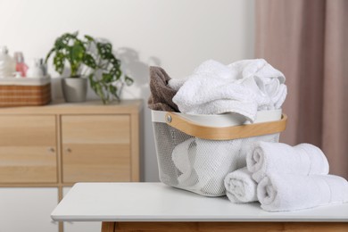 Laundry basket overfilled with clothes near rolled towels on white table indoors. Space for text