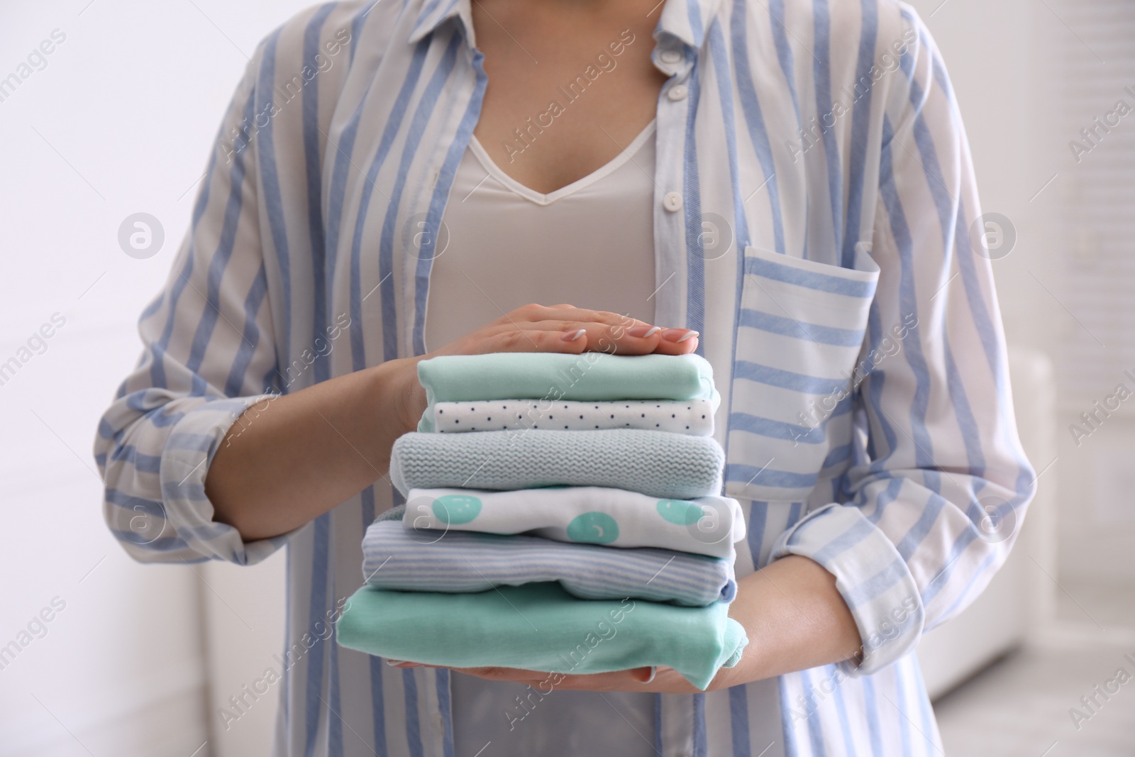 Photo of Woman holding stack of baby's clothes indoors, closeup