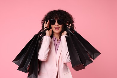 Happy young woman with shopping bags and stylish sunglasses on pink background