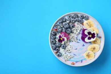 Delicious smoothie bowl with fresh fruits, blueberries and flowers on light blue background, top view. Space for text