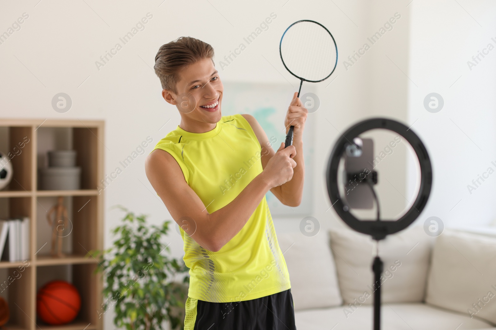 Photo of Smiling sports blogger holding badminton racket while streaming online fitness lesson with smartphone at home