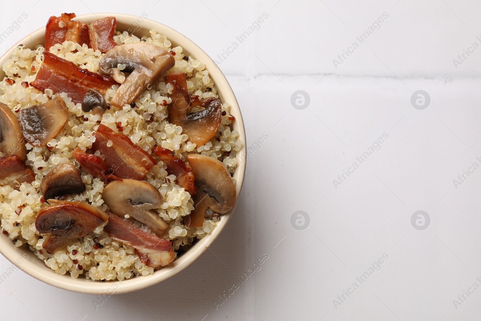 Photo of Tasty quinoa porridge with fried bacon and mushrooms in bowl on white tiled table, top view. Space for text