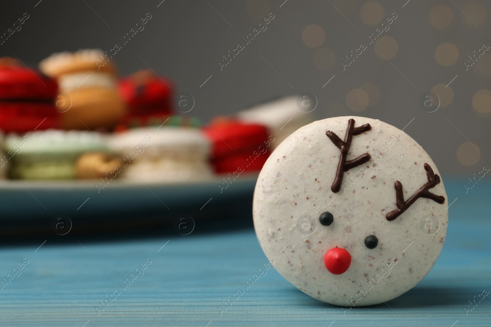 Photo of Tasty reindeer Christmas macaron on light blue table against blurred festive lights, space for text