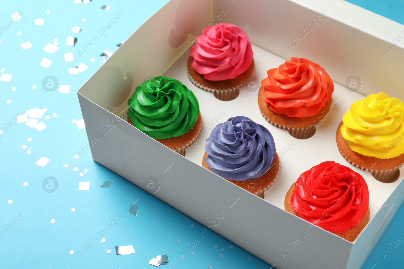 Photo of Box with delicious colorful cupcakes and confetti on light blue background