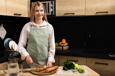 Photo of Young woman in clean apron cutting baguette on wooden table at kitchen