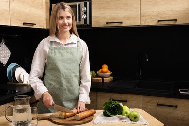 Young woman in clean apron cutting baguette on wooden table at kitchen