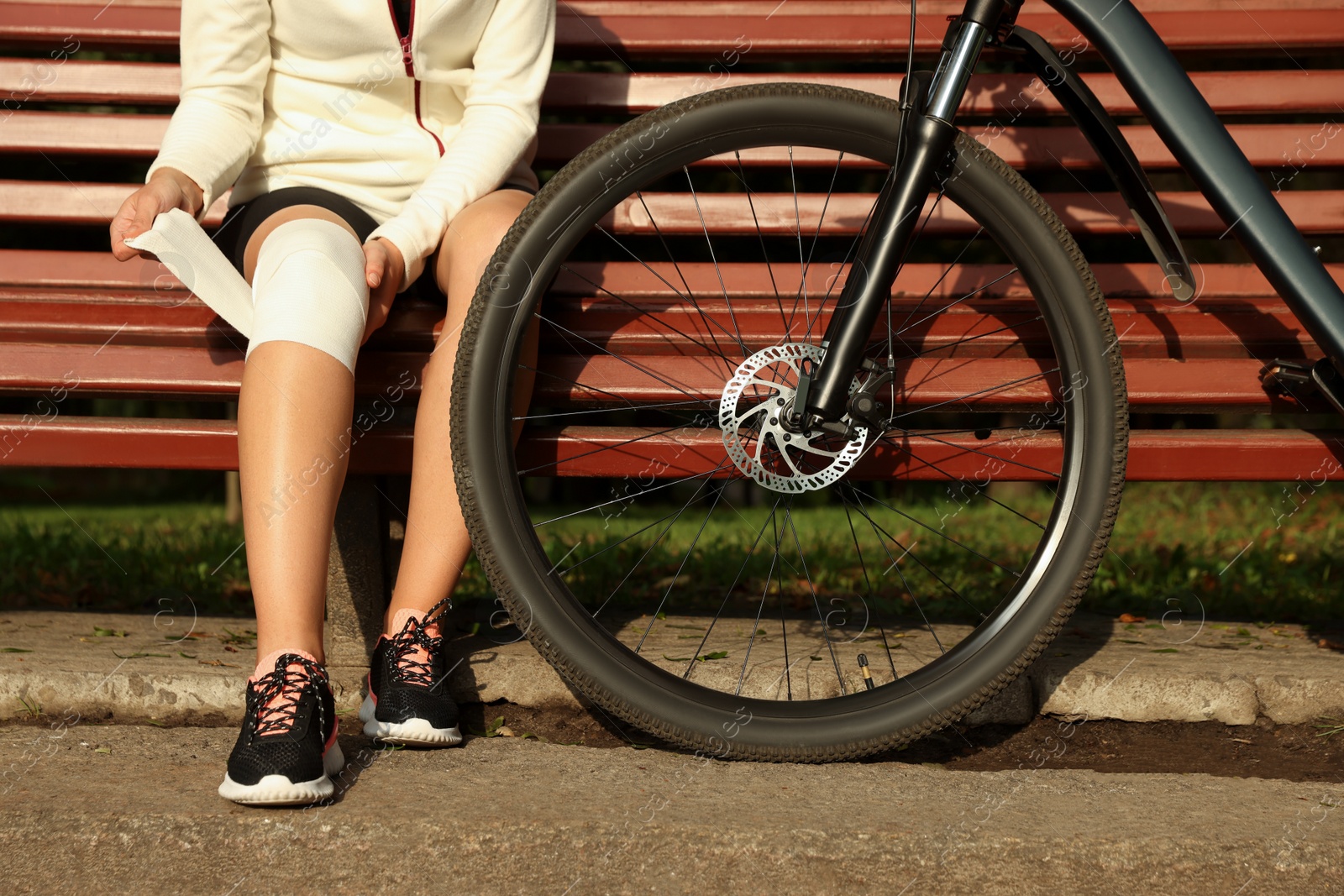 Photo of Young woman applying bandage onto her knee on wooden bench outdoors, closeup