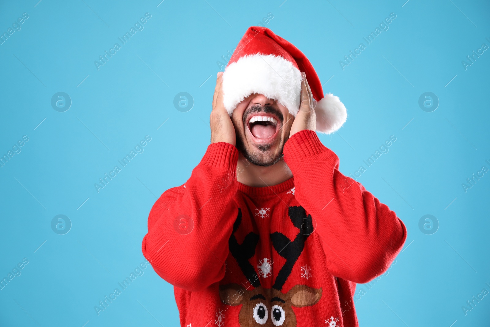 Photo of Emotional young man in Christmas sweater and Santa hat on light blue background