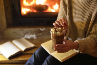 Photo of Woman with glass of hot cocoa and book near fireplace at home, closeup. Space for text