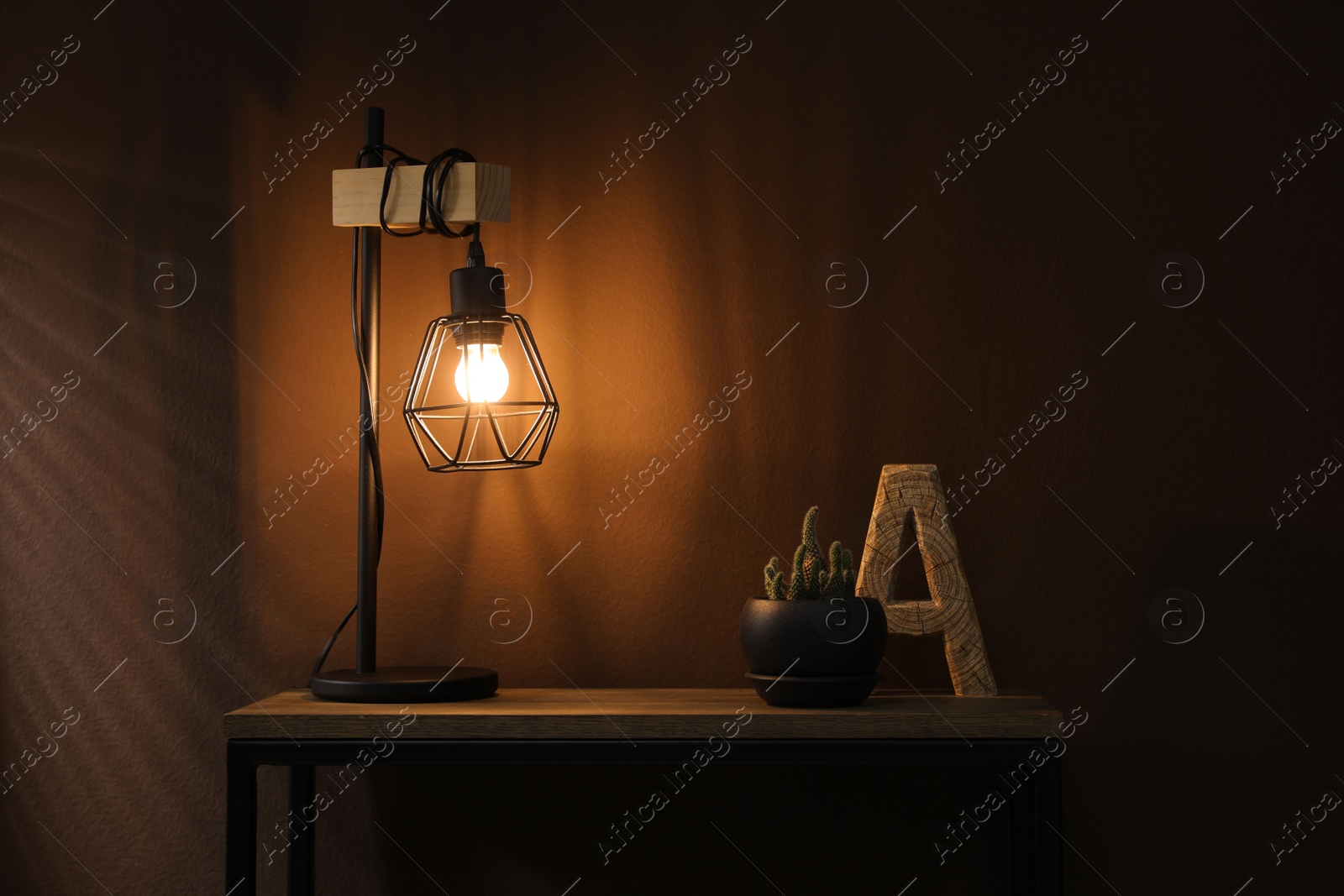 Photo of Stylish lamp, decor and green plant on wooden table near brown wall indoors. Interior design