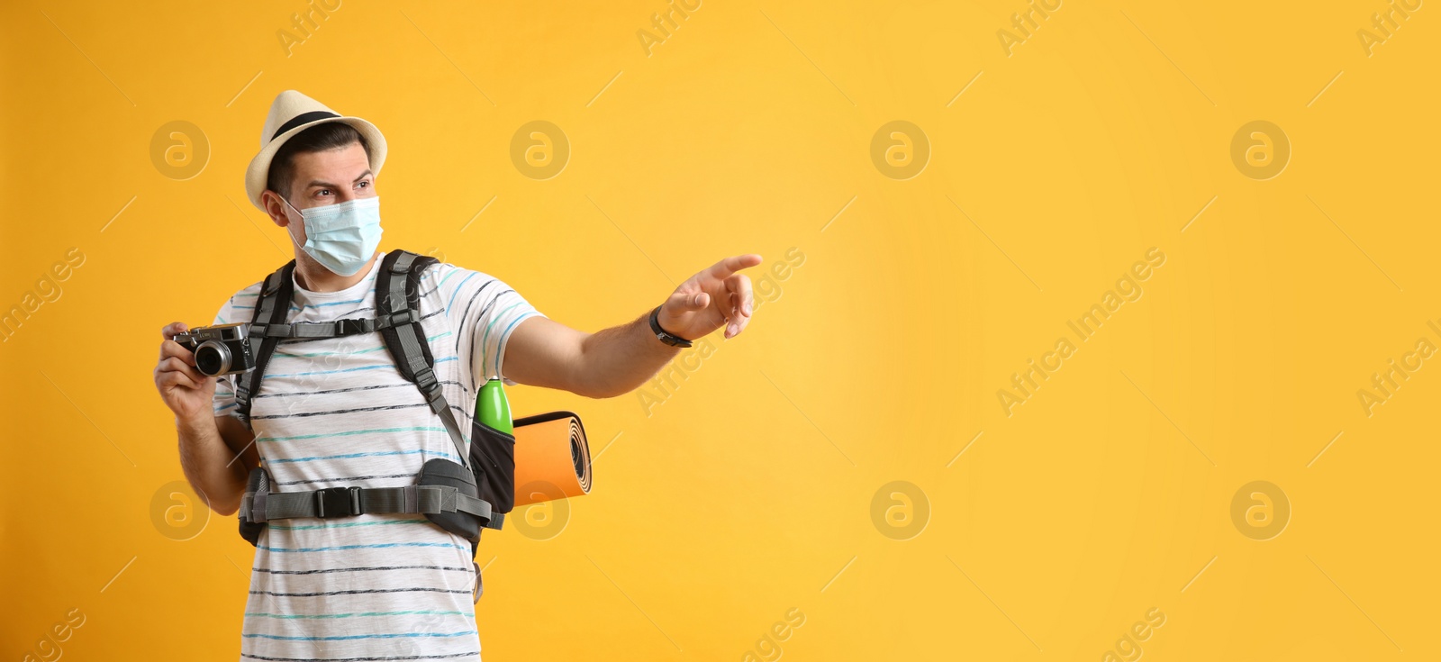 Photo of Male tourist in protective mask with travel backpack and camera on yellow background