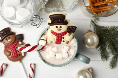 Photo of Flat lay composition with chocolate snowman candy in cup of drink on white wooden table