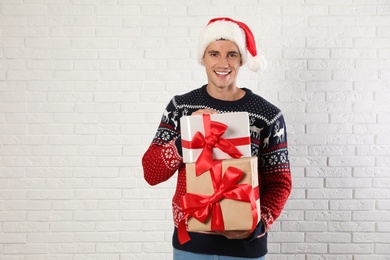 Happy man in Christmas sweater and Santa hat holding gift boxes near white brick wall