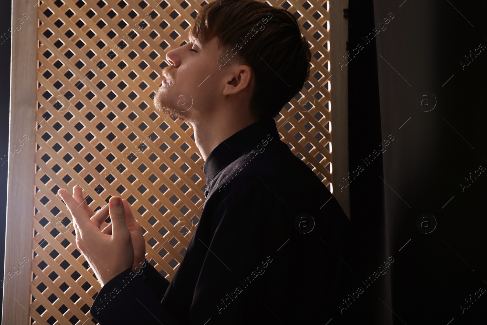 Photo of Catholic priest praying near wooden window in confessional booth