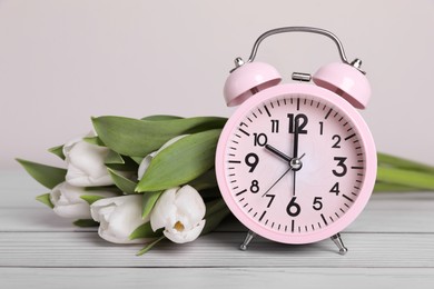 Pink alarm clock and beautiful tulips on white wooden table against light background, closeup. Spring time