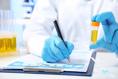 Laboratory assistant with urine sample for analysis writing medical report at table, closeup