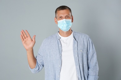 Photo of Man in protective mask showing hello gesture on grey background. Keeping social distance during coronavirus pandemic