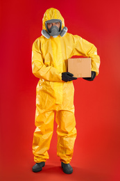 Photo of Man wearing chemical protective suit with cardboard box on red background. Prevention of virus spread