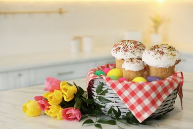 Photo of Basket with traditional Easter cakes, dyed eggs and flowers on table indoors, space for text