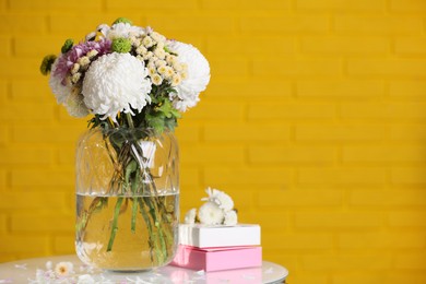 Photo of Bouquet of beautiful chrysanthemum flowers and boxes on white table against yellow brick wall, space for text