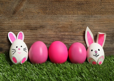 Two white eggs as Easter bunnies among others on green grass against wooden background