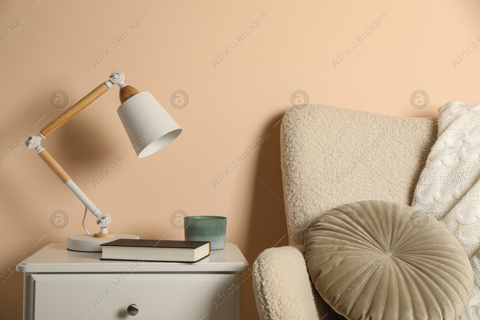 Photo of Stylish modern desk lamp, book and candle on white cabinet near soft armchair indoors