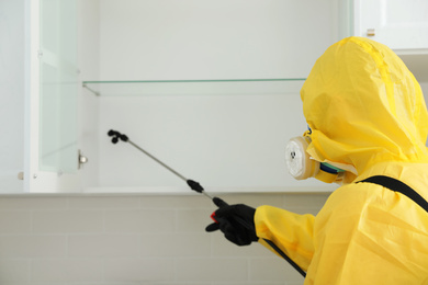 Pest control worker in protective suit spraying insecticide on furniture indoors