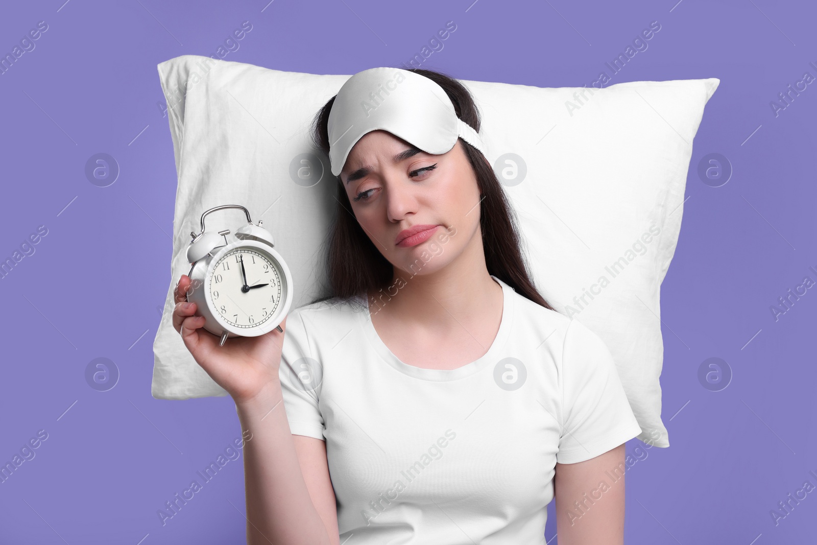 Photo of Tired young woman with sleep mask, alarm clock and pillow on purple background. Insomnia problem