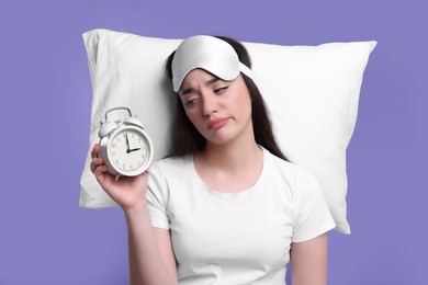 Photo of Tired young woman with sleep mask, alarm clock and pillow on purple background. Insomnia problem