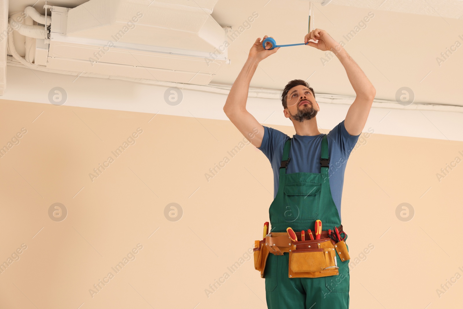 Photo of Electrician in uniform with insulating tape repairing ceiling wiring indoors. Space for text