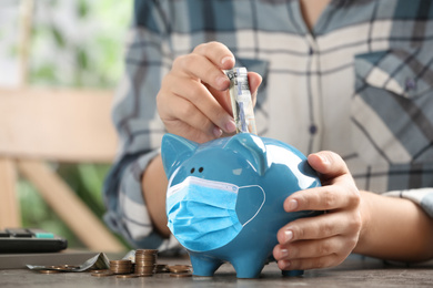 Image of Woman putting money into piggy bank with face mask at table, closeup